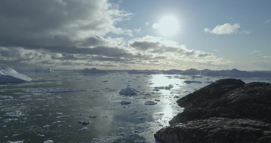 An aerial of the arctic ocean with icebergs and glaciers in it with the sun reflecting on the water