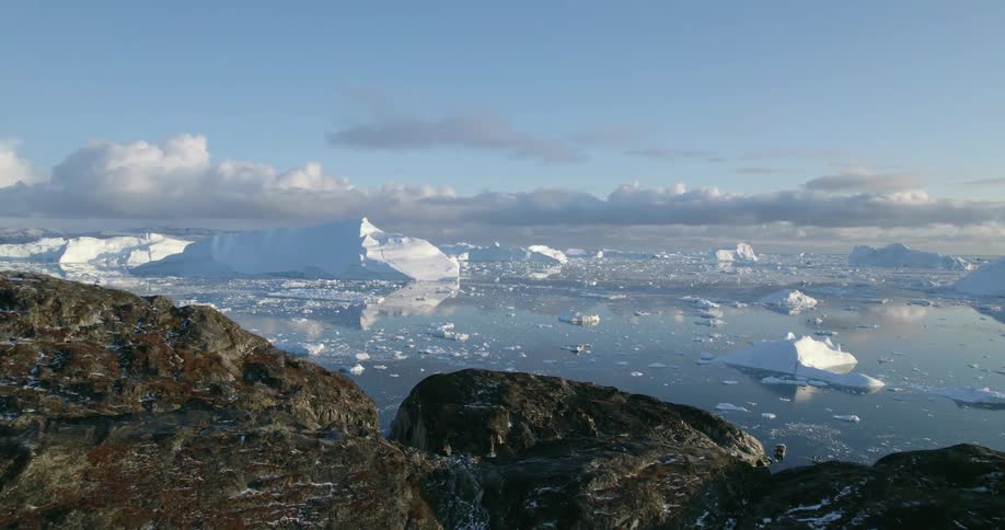 Looking over a mountain as the sun shines brightly as pieces of ice float among icebergs in the water under a clear blue sky