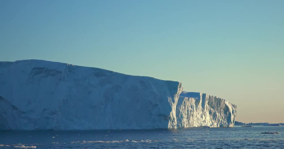 The sun shines brightly as pieces of ice float among icebergs in the water under a clear blue sky