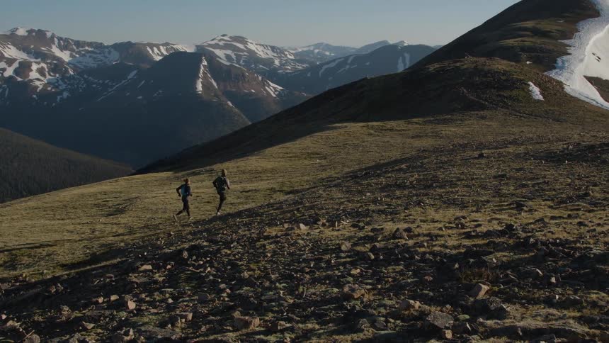 Two people running across hilly grasslands toward a mountaintop with snow on it