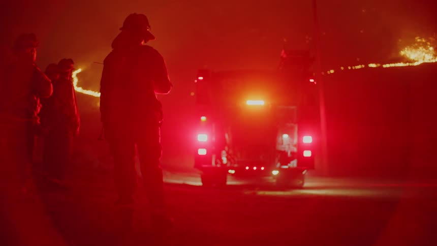Firefighters stand a distance from wildfire as flashing lights on fire engine color area red