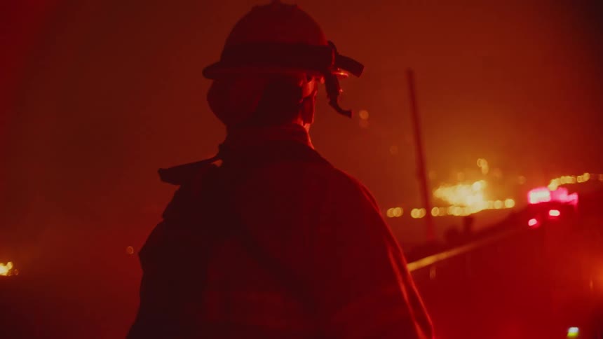 Firefighter stands looking past flashing red lights toward extensive wildfire at night