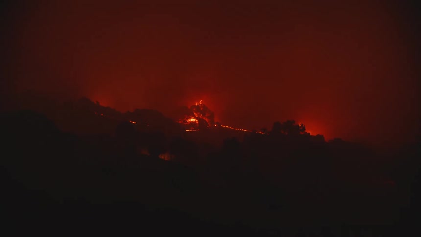 A wildfire is burning woods in the distance while filling the sky with flames and smoke