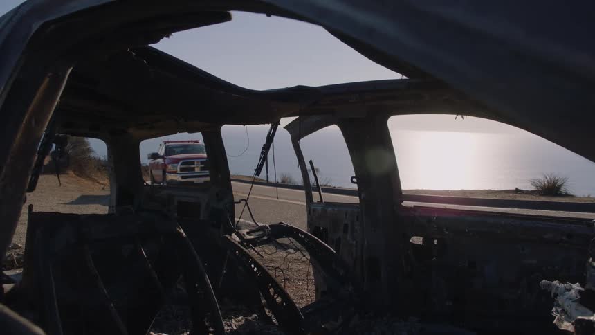 An old burnt car on the side of the road as a truck drives by slowly