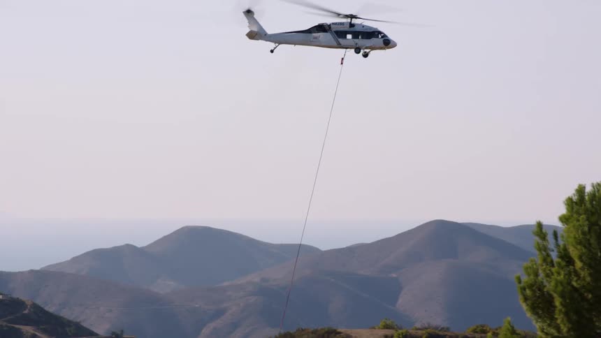 Helicopter hauls helicopter water bucket across field with mountains in the background