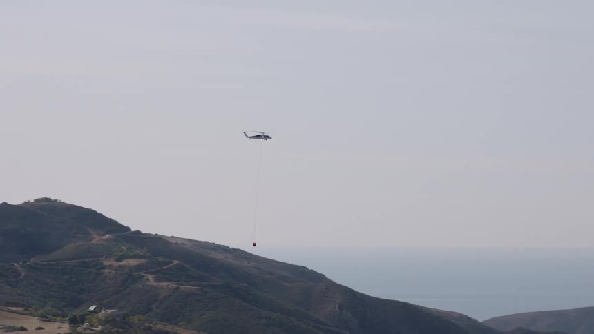 A helicopter carries equipment on a rope ladder over a mountain range