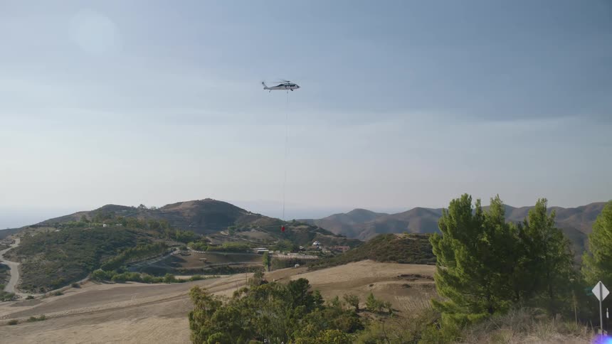 A helicopter lowers itself into a dry grassy plain between tress and distant hill peaks