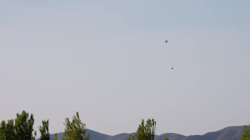 A helicopter carrying a water bucket is flying over mountains