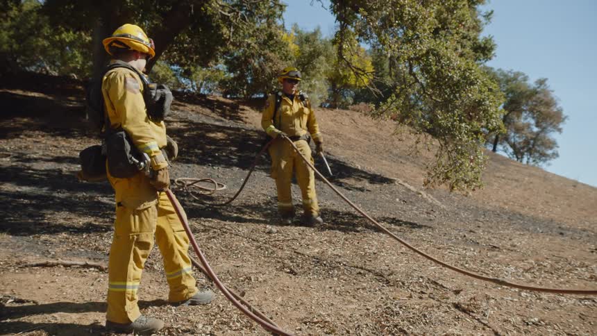 Two firemen are holding a hose as they walk on a hillside near trees and the sun shines brightly upon them