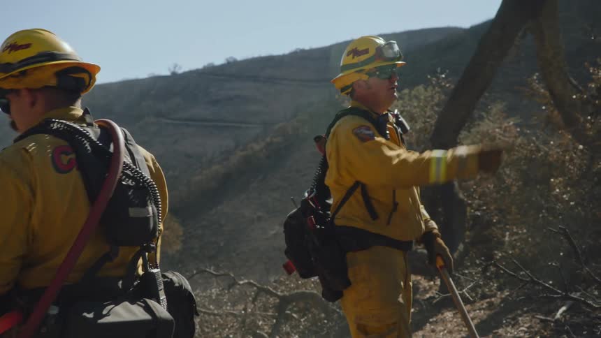 Two firefighters in yellow suits dig out plant life in a brown canyon to prevent forest fires