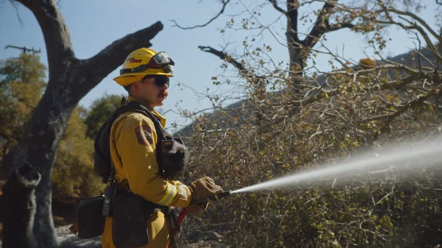A firefighter sprays water in a forest as other firefighters walk around