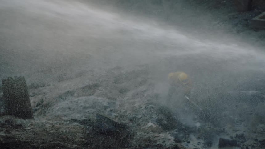 A fireman is turning over soil and rocks and water is spraying over the area while fighting a forest fire