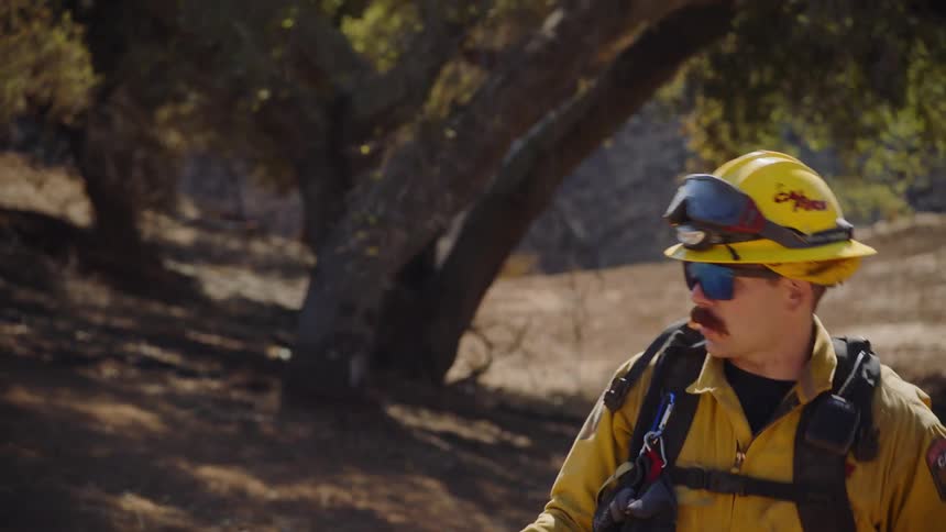 A firefighter wearing yellow gear walks downhill with a hose