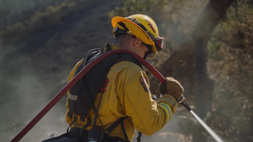 A fireman is standing outside holding a hose over his spraying water on the ground as smoke fills the air