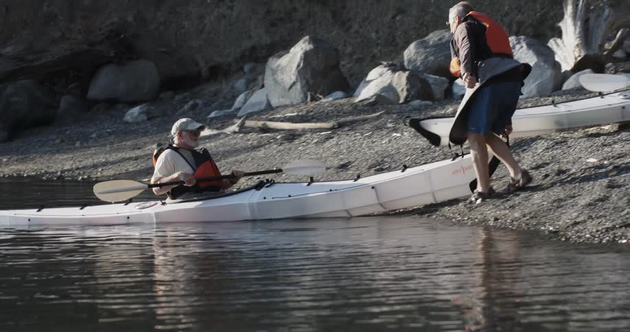 An older man on the beach helps an older man in a kayak bring his kayak to shore