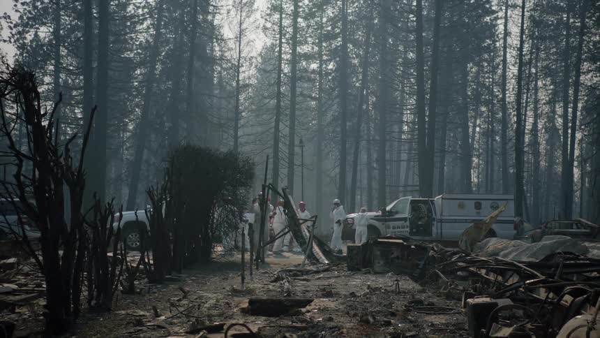 People wearing white jumpsuits and helmets are walking in the smoke filled woods around debris and ruins where a house once stood and trucks are parked nearby