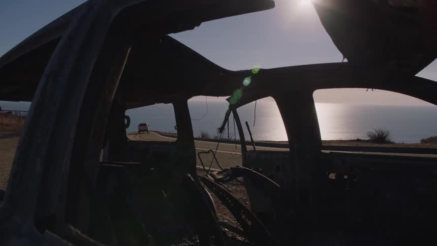 Vehicle drives along coastal highway passing burned car wreckage