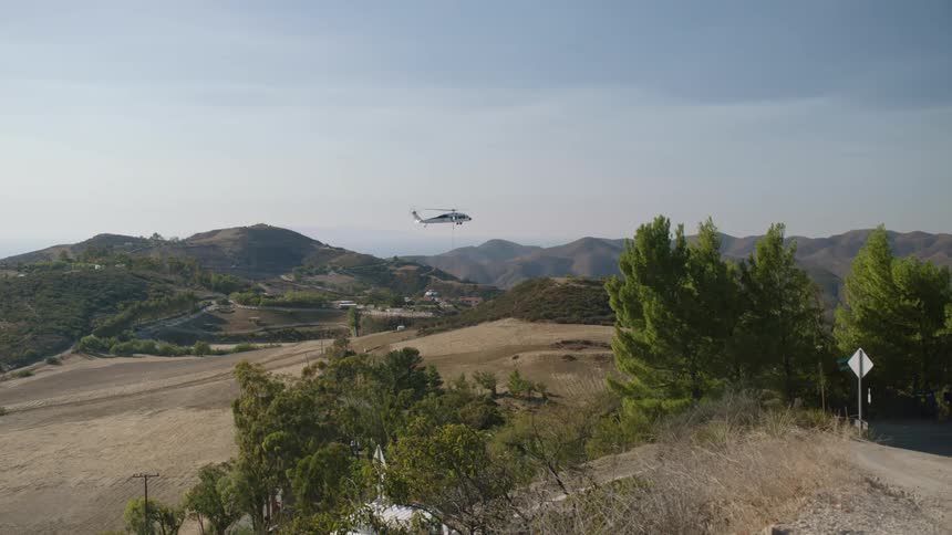 A helicopter is hoovering in the sky near mountains as a truck travels around the bend on a mountain road