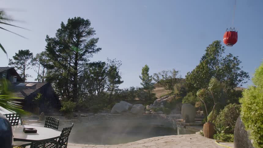 Helicopter removes water from a pool next to a house and some trees and carries it away on a sunny day