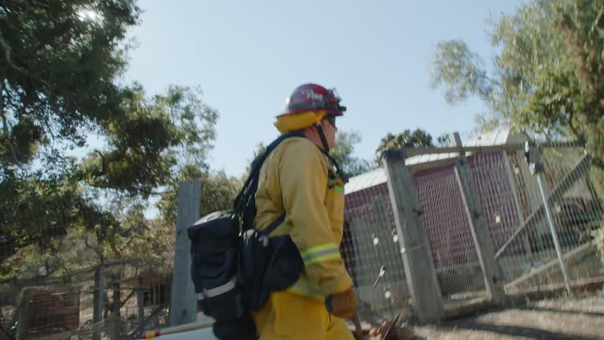 A fireman wearing a backpack is pulling a hose as he walks along a fence