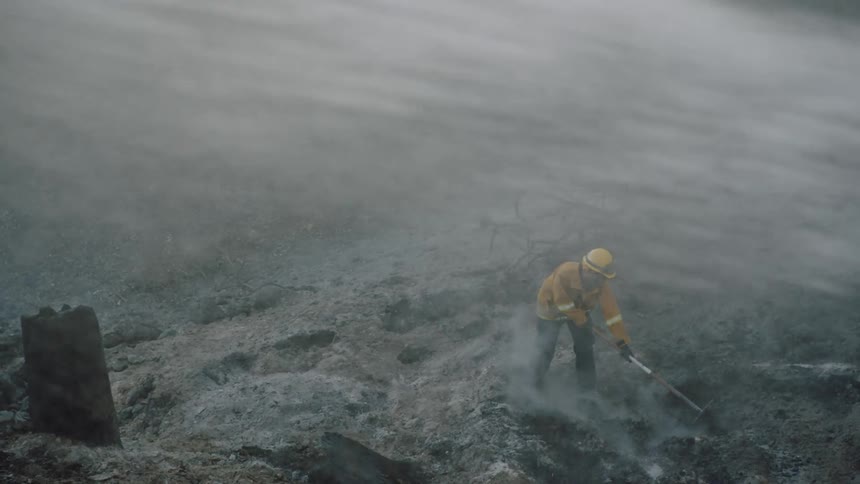 Firefighters wearing yellow protective gear rakes the ashes on the ground after a forest fire