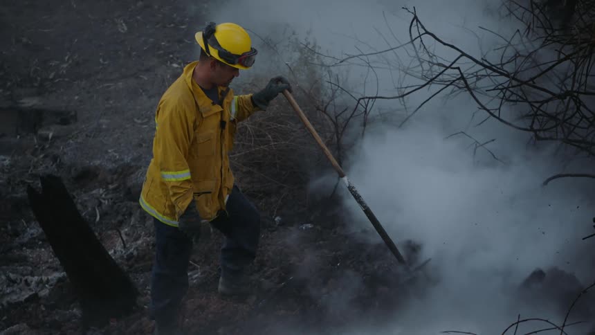 A firefighter stands in the aftermath of a major disaster while smoke clouds rise from the ground and water sprays on it