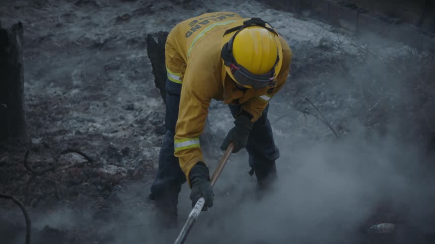 The aftermath of a fire is shown while a firefighter uses a fire broom to rake gravel near something expelling water