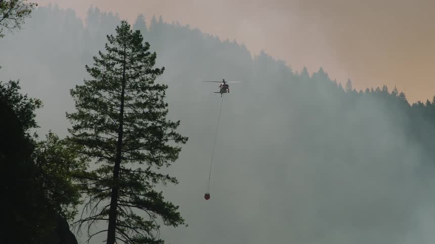 A helicopter is flying through the smoke of a forest fire carrying a bucket of water underneath it on a rope