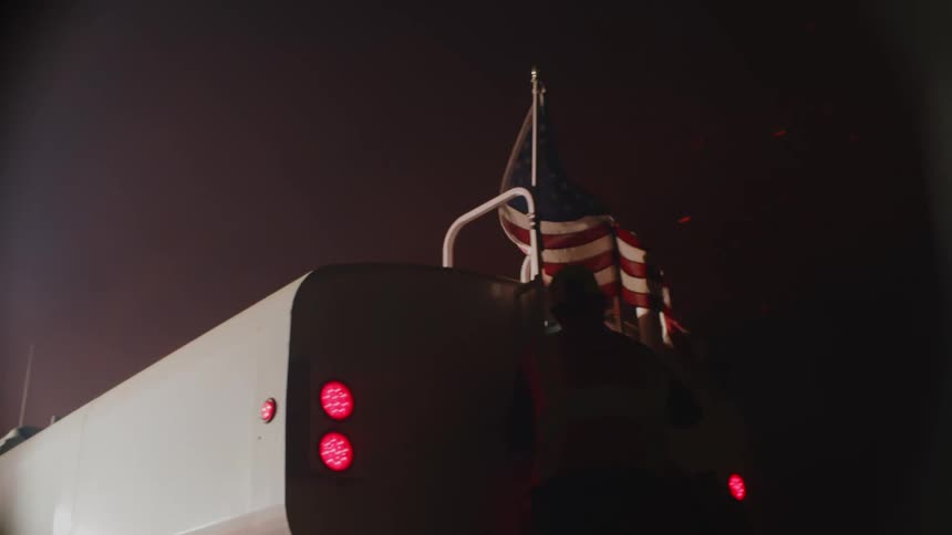 An american flag flies on the back of a truck as it waves in the wind and a man is standing on the back