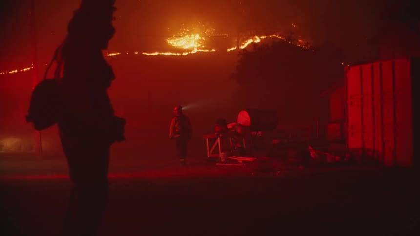 Firefighters stand in lot lit by red flashing light at night near burning slope