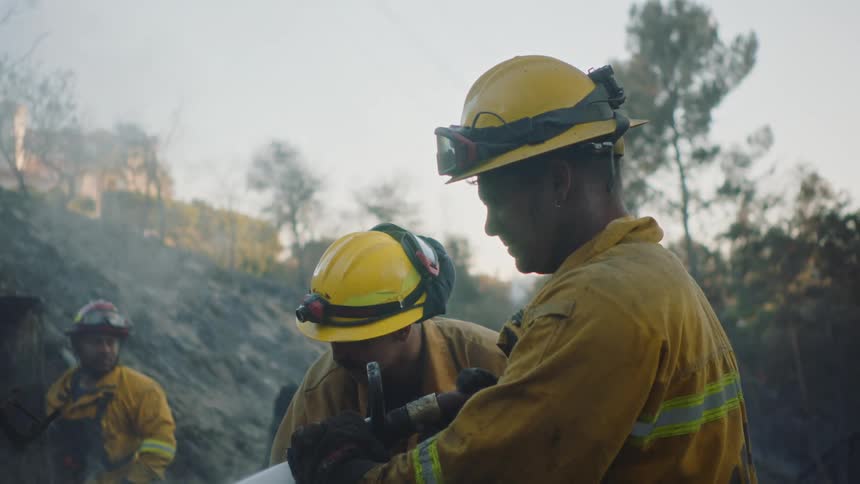 Three firemen standing spraying a hose at the ground while another man rakes the groudn