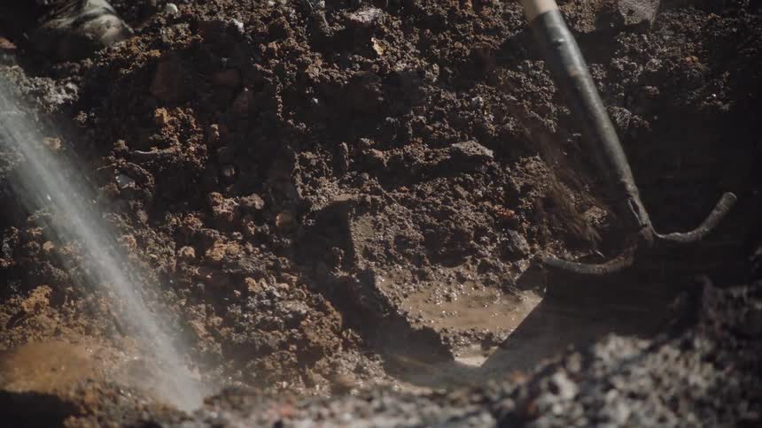 A close up of muddy soil as a person sprays water into the whole and another person uses a tool to scrap the mud