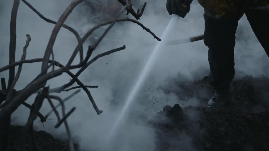 A firefighter spraying water out of a hose at the ground as the ground steams and smokes