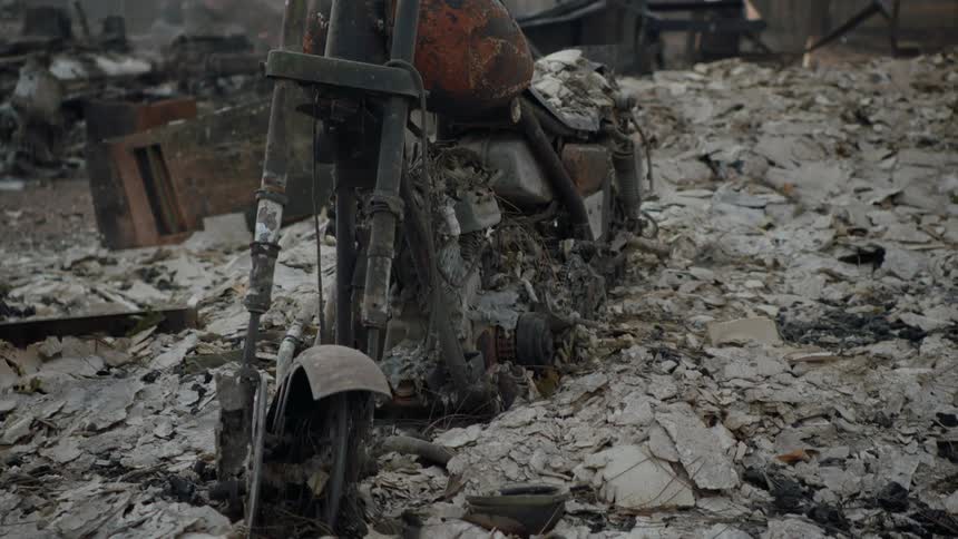 A burned motorcycle sits in ruin amidst rubble in the middle of a forest after a natural disaster