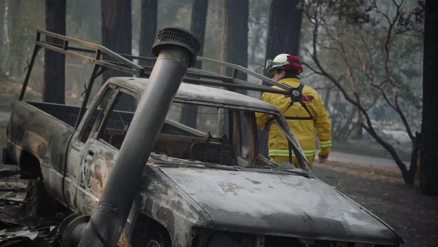 A fireman looks in the window of a burned out truck in the woods and he walks away as smoke fills the air