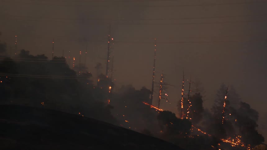 Fire and flames climb up trees on a mountain in a smoke filled sky as the wildfire spreads