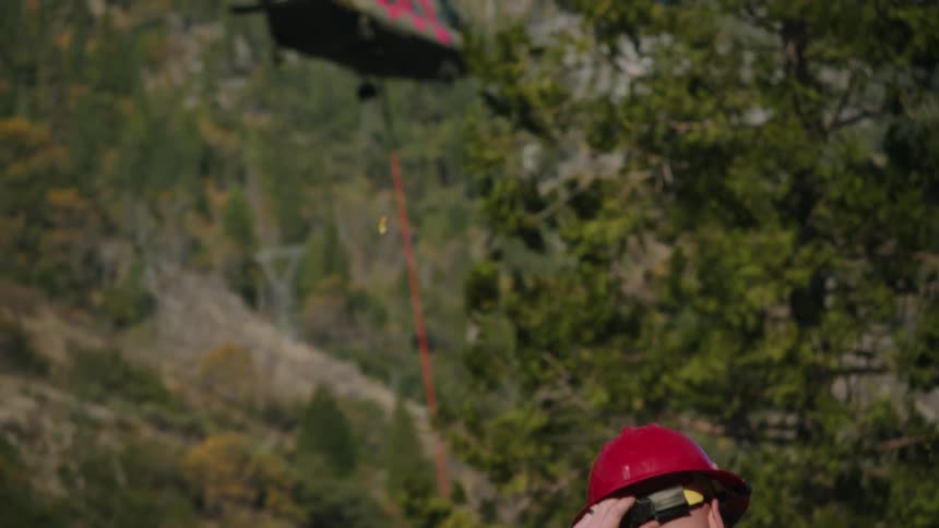 Firefighter wearing yellow protective gear and red helmet waits for a helicopter over the mountains