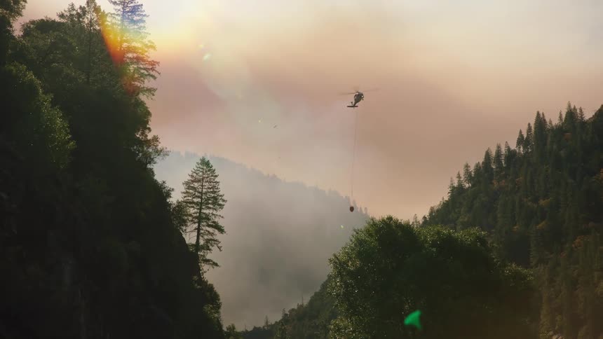 A helicopter spraying a forest fire below it as smoke billows from the ground