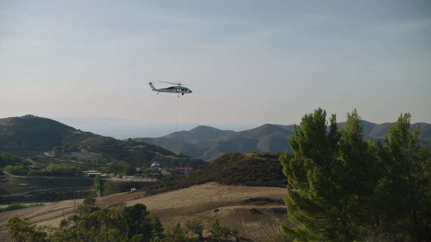 A helicopter is hoovering in the sky as it pulls a bucket of water and flies away with mountains in the distance