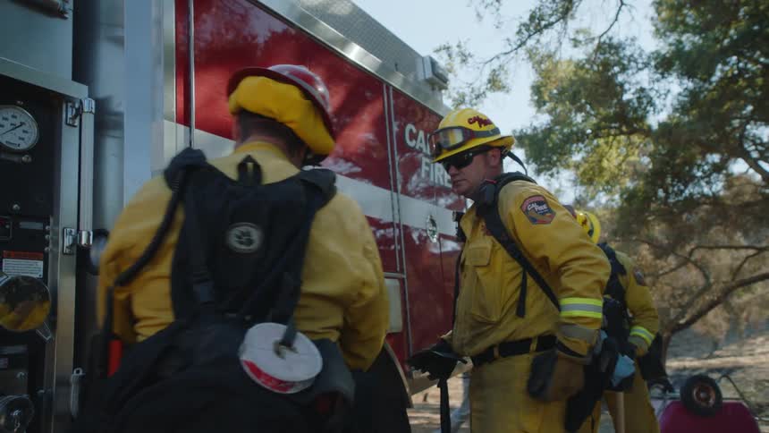 Three firefighters putting equipment on and closing the firetruck as a person in a fireman coat and wearing film equipment walks by