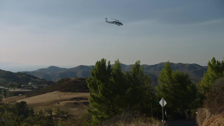 Helicopter lifts large bucket and flies past chain linked fence and discarded fallen stuffed toys