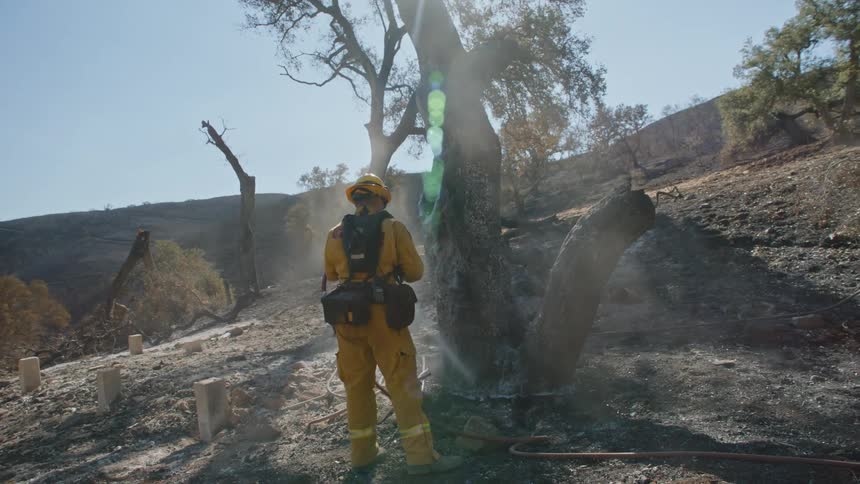 Firefighter stands and hoses down charred area as another firefighter walks down slope toward him