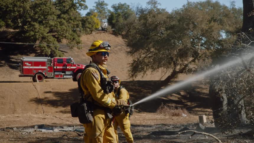 A firefighter puts out a forest fire using a water hose on a sunny day while another firefighter walks by and watches