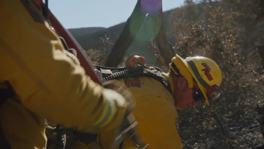 Two firefighters are working outside as one is digging and the other is holding a hose and spraying water on the ground