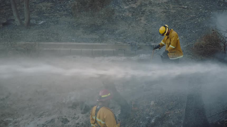 Two firefighters stand on slope as stream of water from hose flows onto hot smoking ground