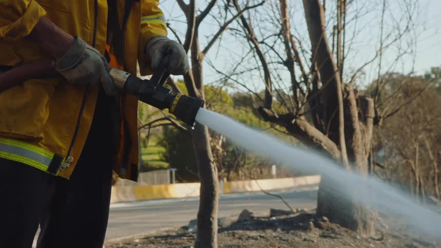 A firefighter in a yellow coat uses a highpowered hose to spray water into dry dirt trench