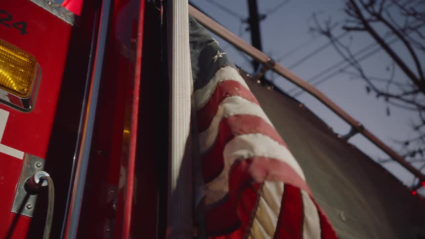 A dirty us flag hangs off the side of a fire truck with flashing lights outdoors at dusk