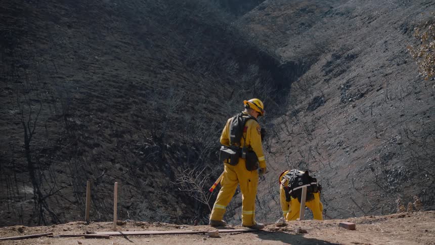 A firefighter pokes ashes by a cliff another firefighter watches and then walks away