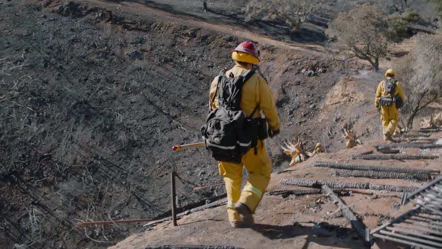 Two firefighters walk along a cliff in a canyon next to a the roof of a collapsed dwelling