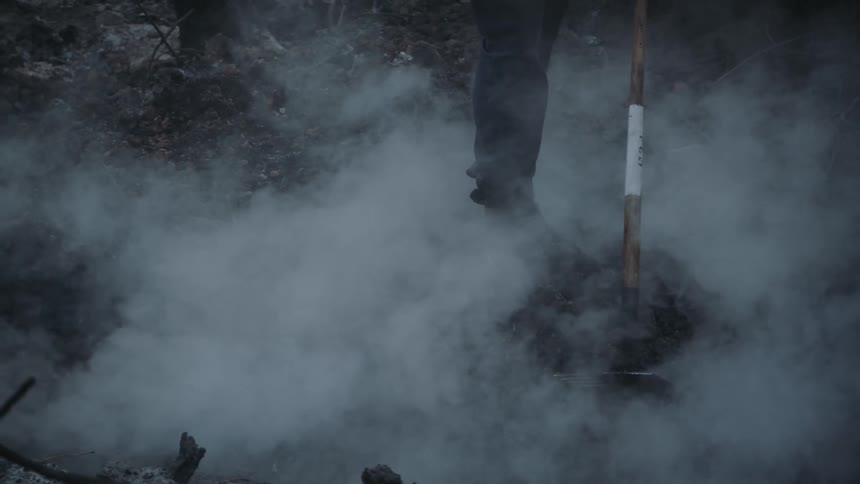 Man standing amidst the smoldering ashes of a forest fire while leaning onto a rake handle